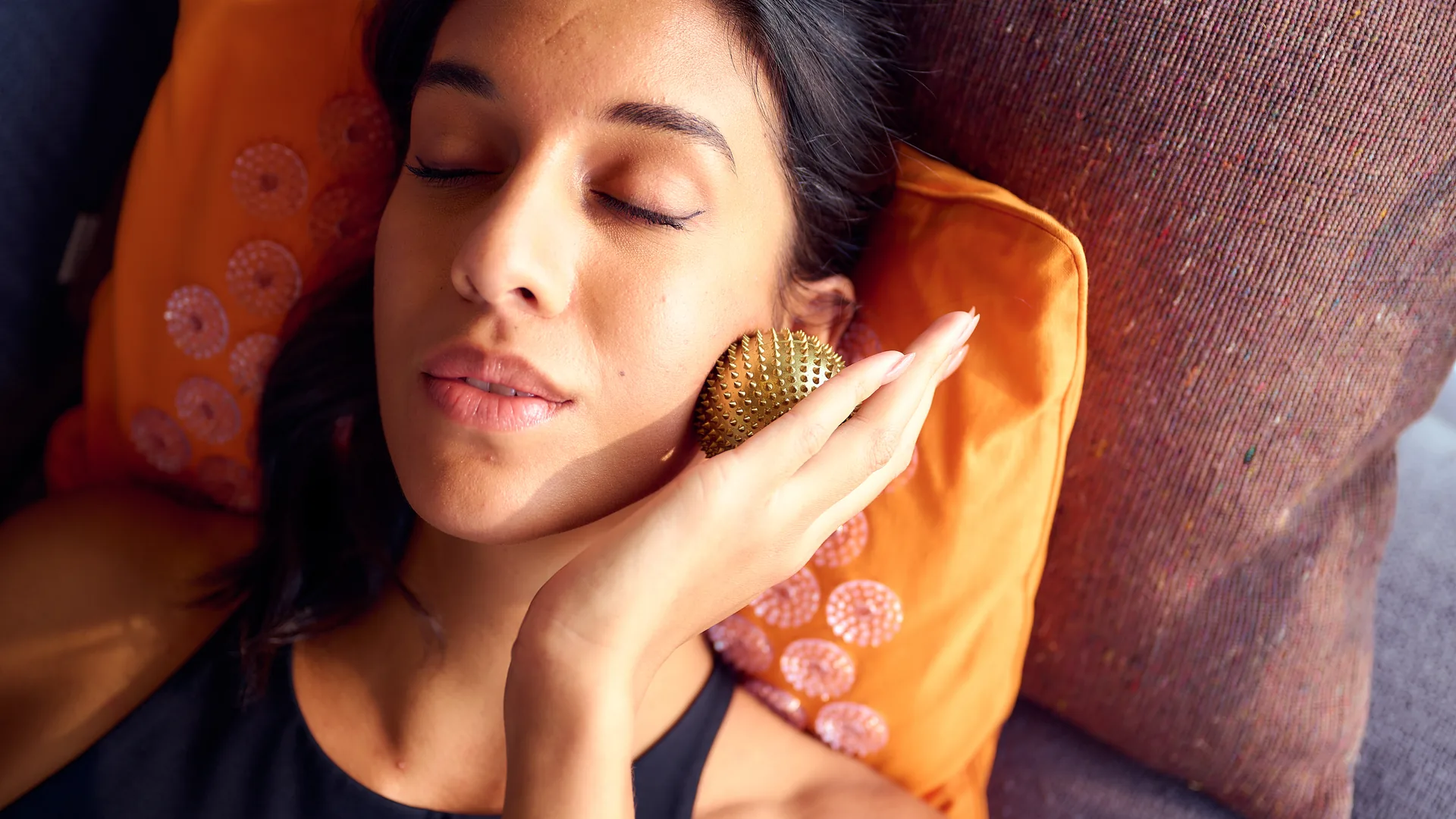 A teacher reclines on a Shakti acupressure pillow, gold Wonderball resting against her jaw — tension release and recovery